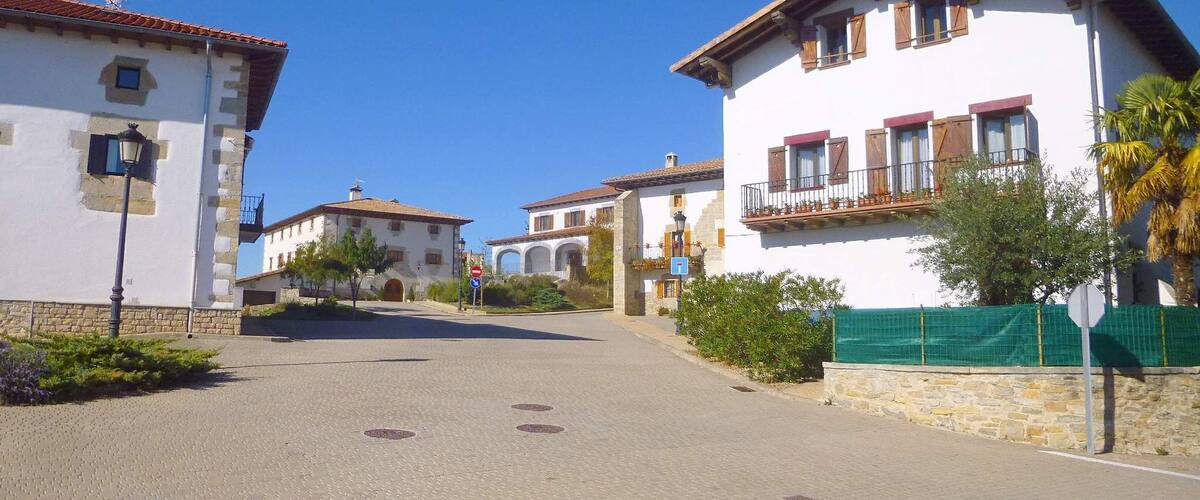 A sunny street in a European village with white buildings, a clear blue sky, and a green fence.