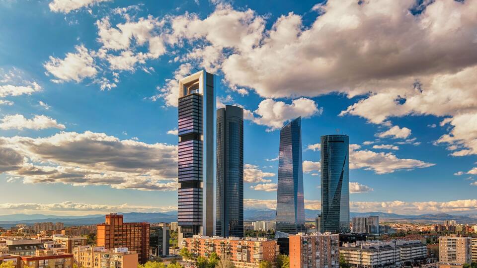 Un paisaje urbano con rascacielos modernos y montañas al fondo, bajo un cielo parcialmente nublado.