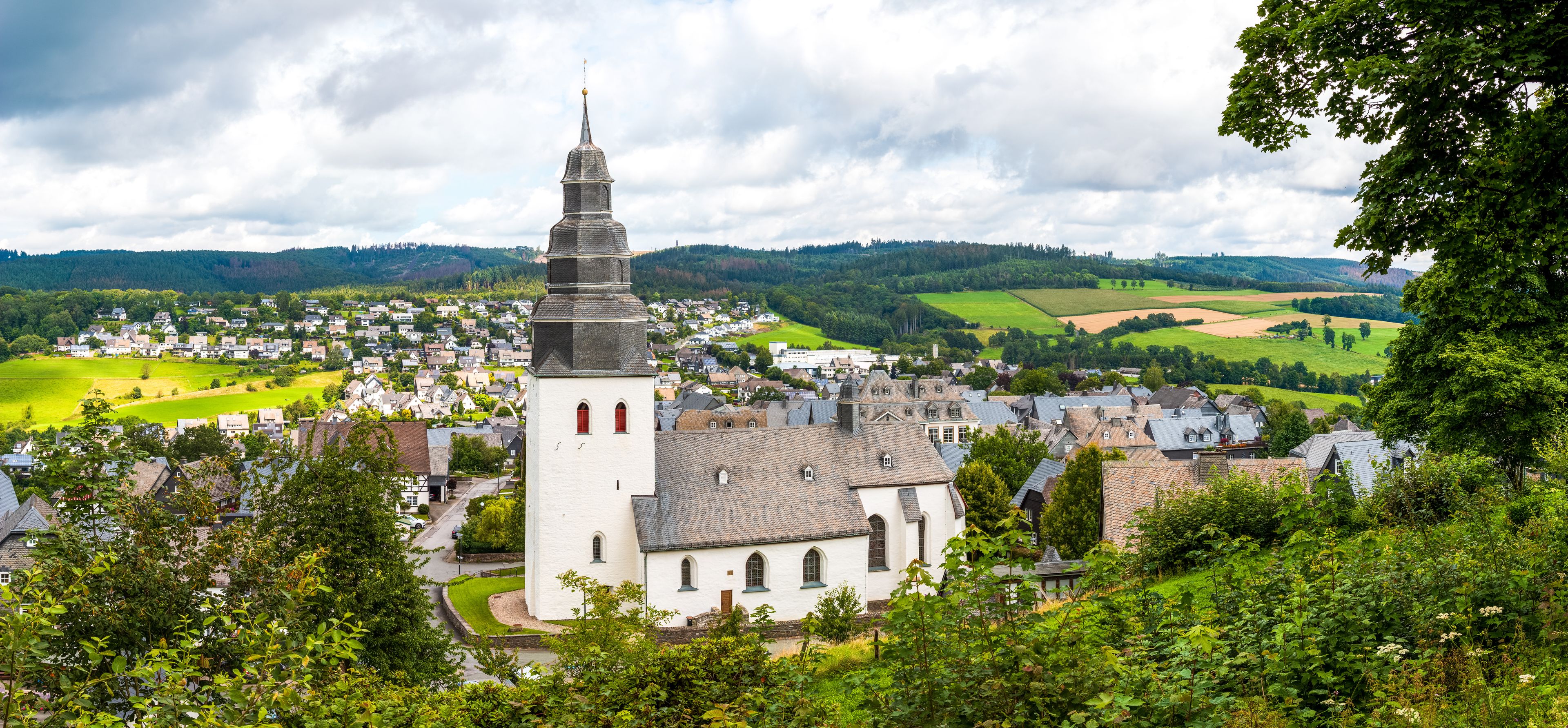Une ville où se dresse une église remarquable, reconnaissable à son clocher élancé, et qui est entourée d’une végétation luxuriante et de collines ondoyantes.