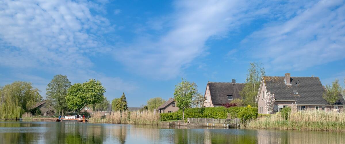 A calm river with a boat, surrounded by trees and houses with thatched roofs.