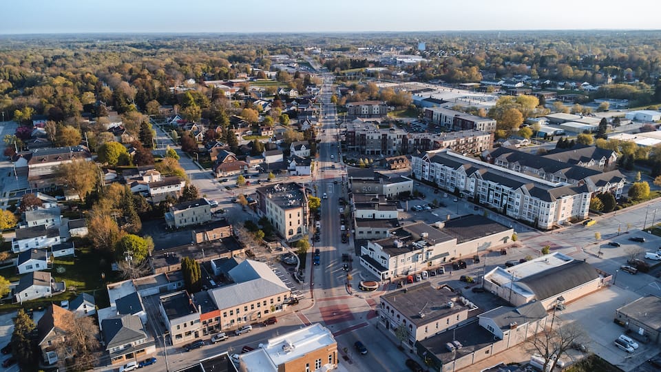 An aerial view of a town with a central street, surrounded by residential and commercial buildings, and a backdrop of trees and open land.