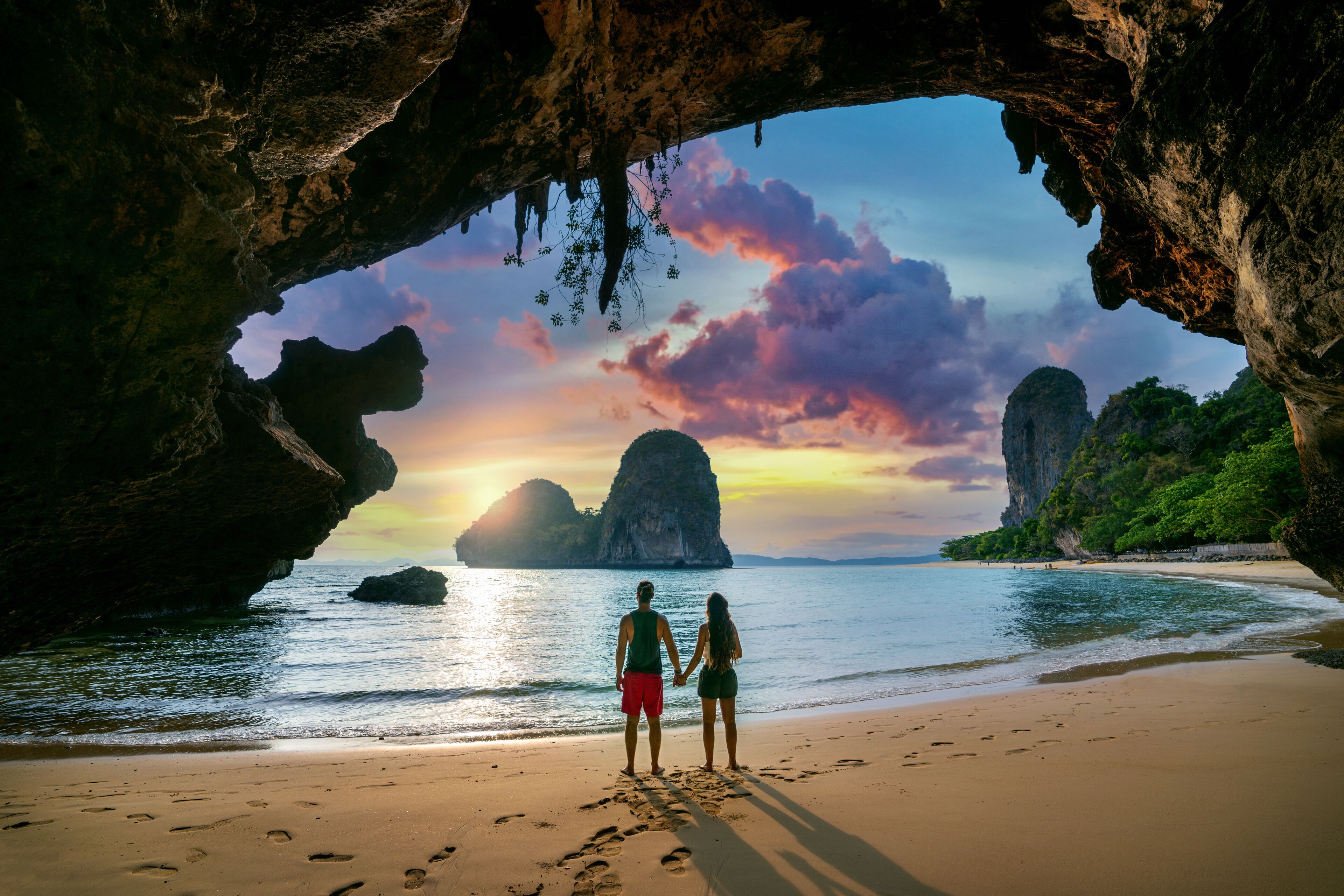 Una pareja de pie en la playa, con una cueva que se abre al mar, un atardecer y formaciones rocosas a lo lejos.