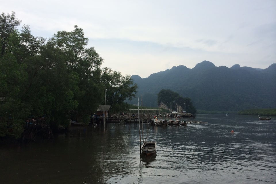 A calm river with boats and a dock on the left, surrounded by lush greenery and mountains in the background.