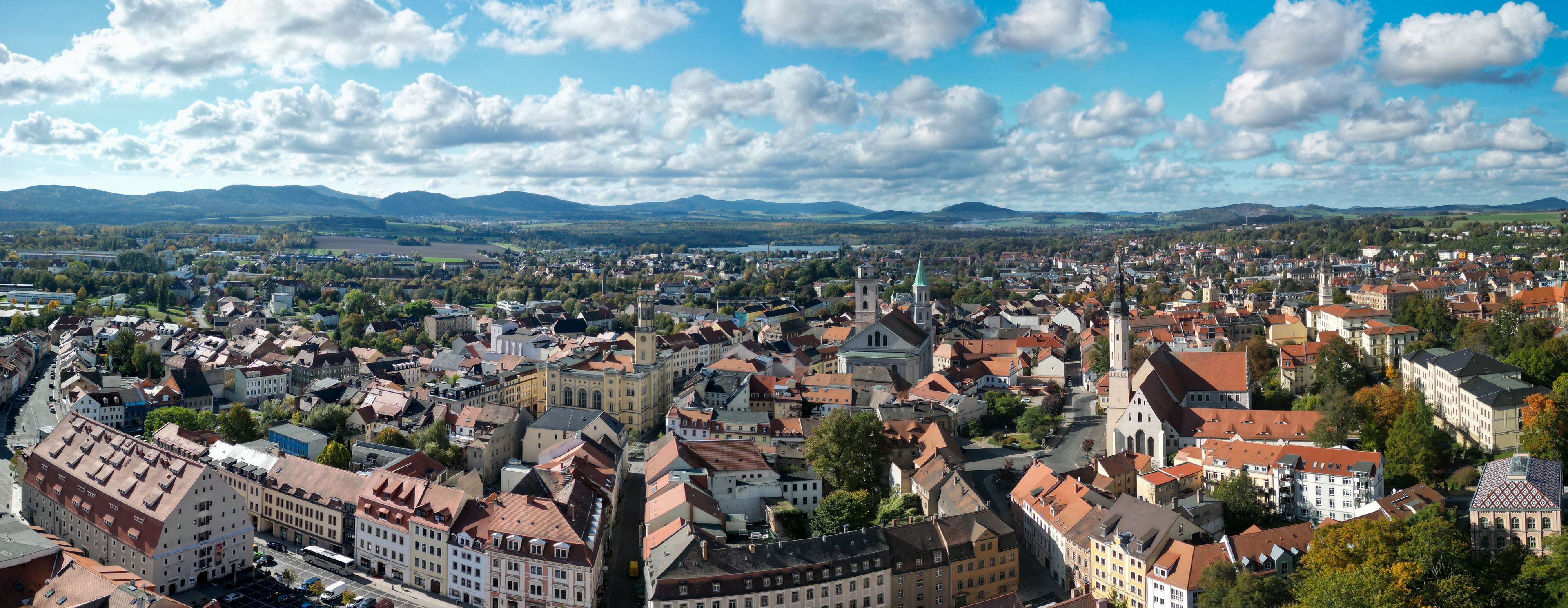 A panoramic view of a historic town with dense architecture, a prominent church tower, and rolling hills in the background.