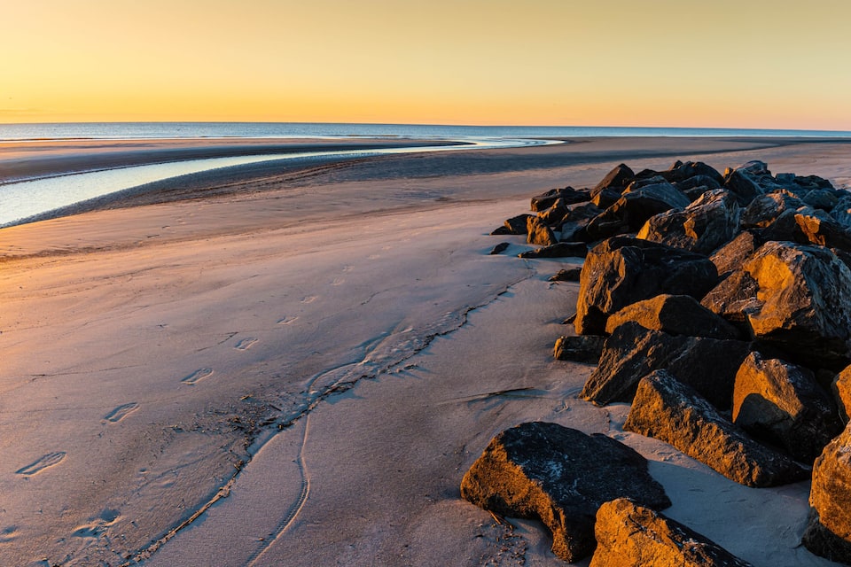 Une plage avec de gros rochers, des empreintes de pas et le soleil se couchant sur l’océan.