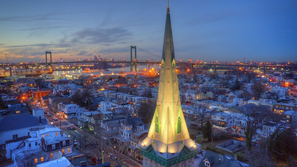 A cityscape with a prominent tower lit up at dusk, a bridge in the background, and snow-covered buildings.