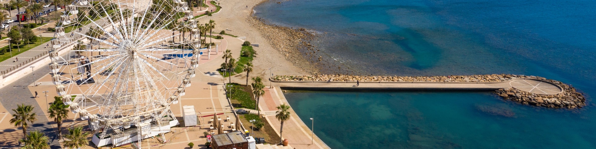 Un paisaje urbano costero con una noria gigante, una playa y un rompeolas rocoso.