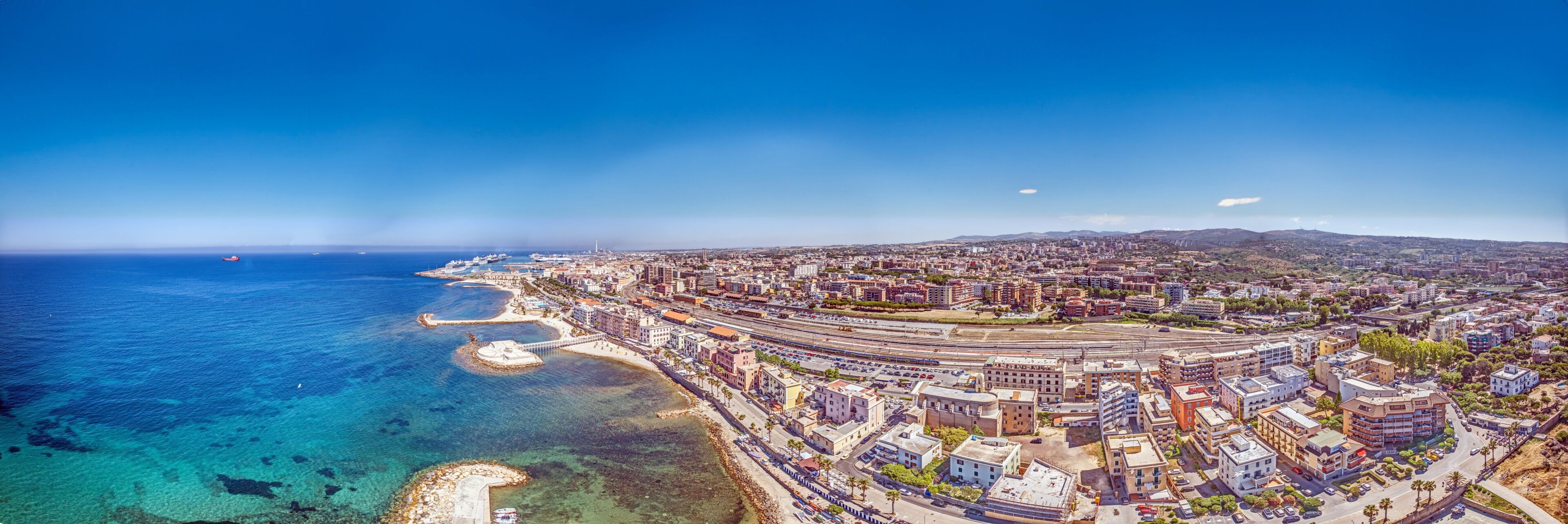 A coastal cityscape with a marina, clear blue sea, and a mix of residential and commercial buildings.