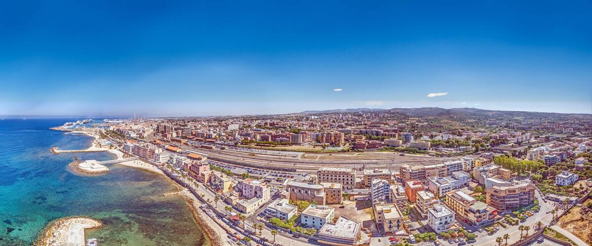 A coastal cityscape with a marina, clear blue sea, and a mix of residential and commercial buildings.
