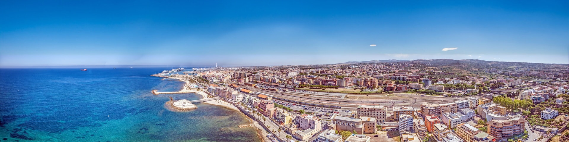 A coastal cityscape with a marina, clear blue sea, and a mix of residential and commercial buildings.