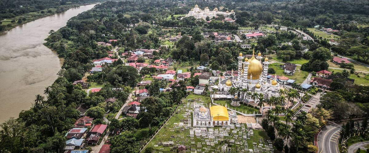 An aerial view of a town with a river, a large white building with golden domes, and surrounding greenery.