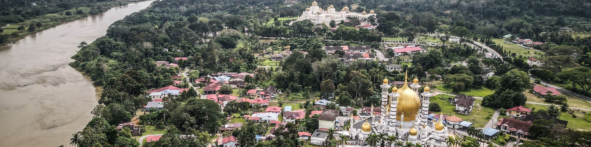 An aerial view of a town with a river, a large white building with golden domes, and surrounding greenery.