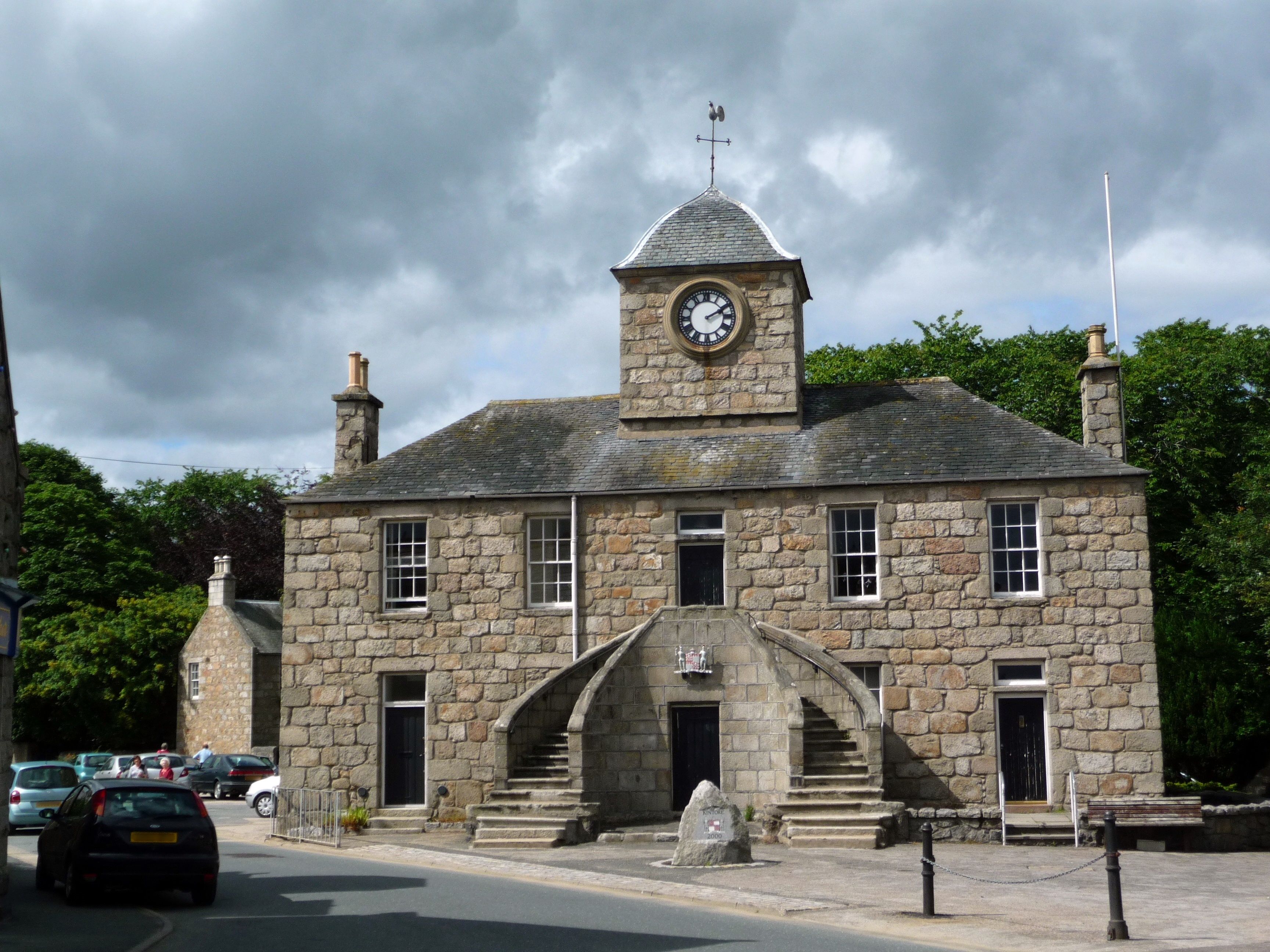 A stone building with a clock tower, a staircase leading to the entrance, and parked cars on the street.