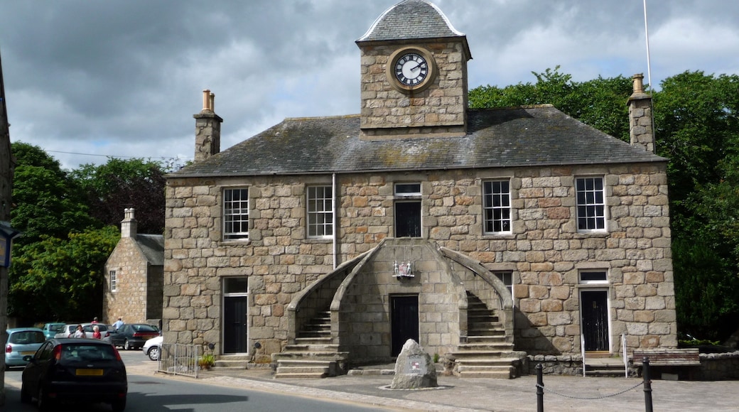 A stone building with a clock tower, a staircase leading to the entrance, and parked cars on the street.
