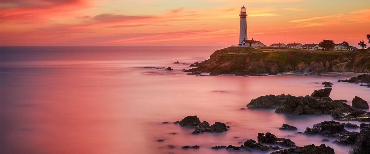 Cena costeira com um farol, linha costeira rochosa e um céu ao pôr do sol.