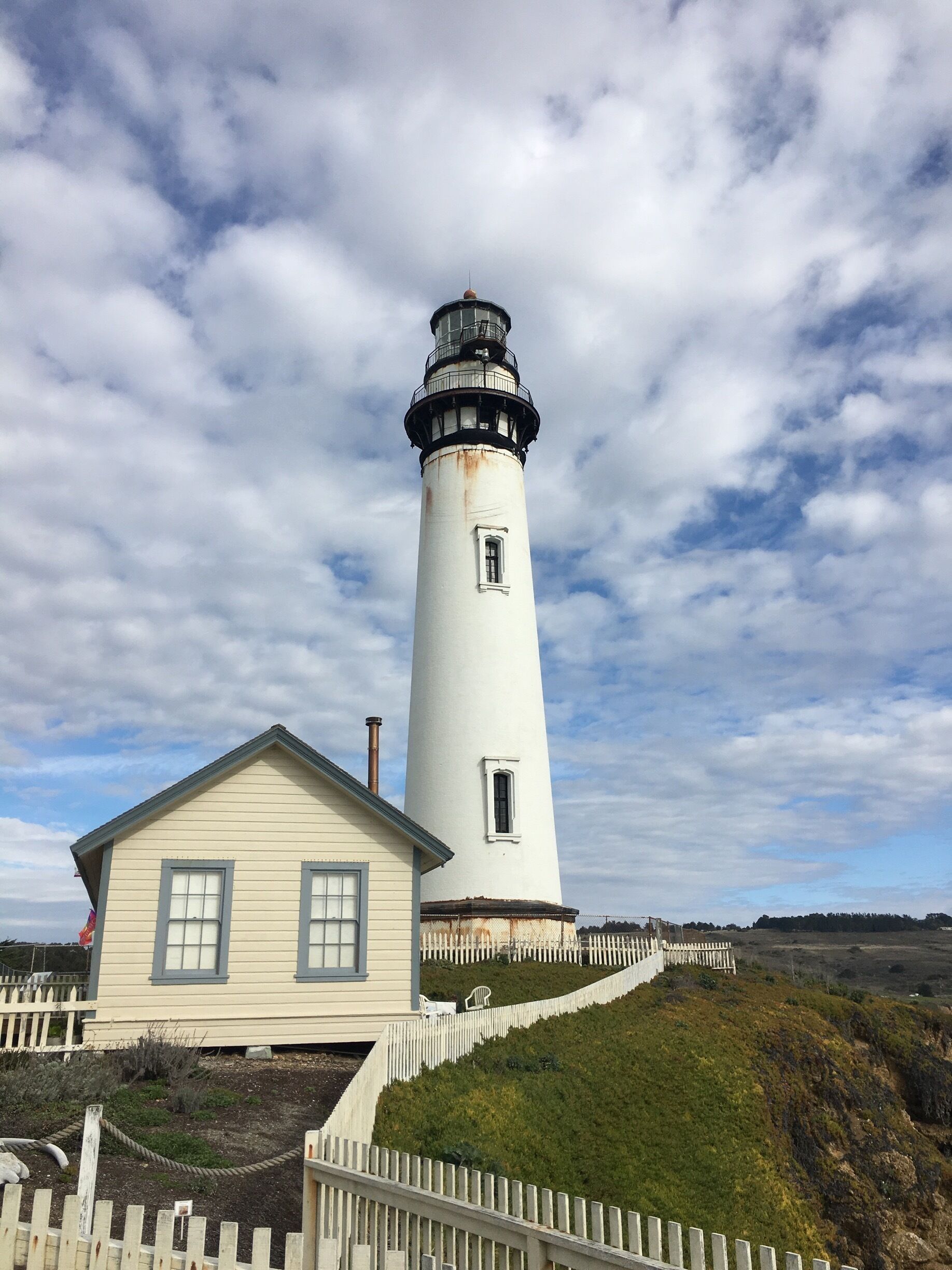 A white lighthouse with a black top, a small white building with a white picket fence, and a cloudy sky.