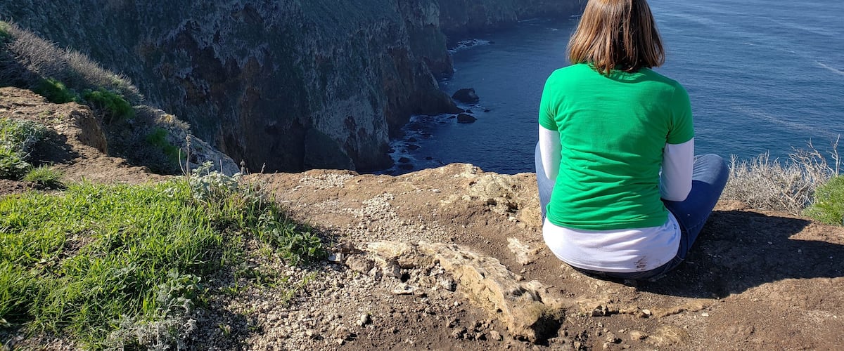 A person sitting on a rocky cliff overlooking a steep cliffside and the ocean.