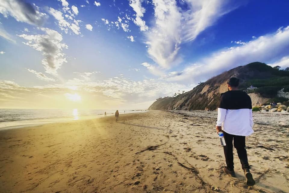Una persona caminando sobre la playa, con huellas, un atardecer y una montaña al fondo.