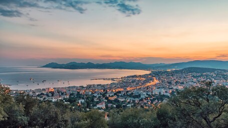 Une vue panoramique d’une ville côtière au coucher du soleil, avec la mer, les montagnes et les bâtiments urbains en arrière-plan.
