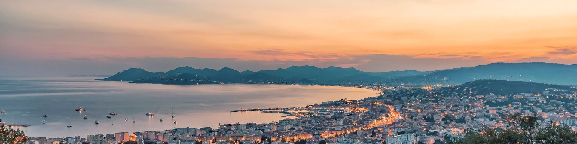 A coastal cityscape at sunset with a view of the sea, mountains, and urban buildings.