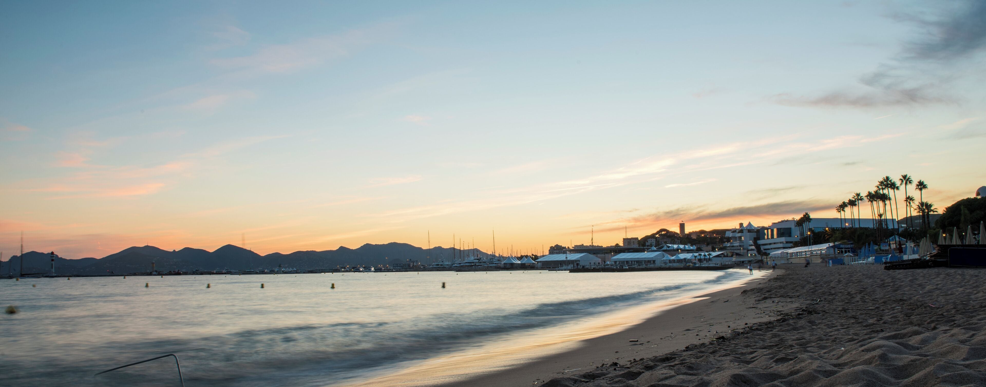 A beach at sunset with calm waters, a lighthouse, and palm trees.