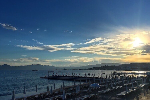 Un coucher de soleil sur la côte, avec une jetée qui s’avance dans la mer, entourée de parasols de plage et de chaises longues.
