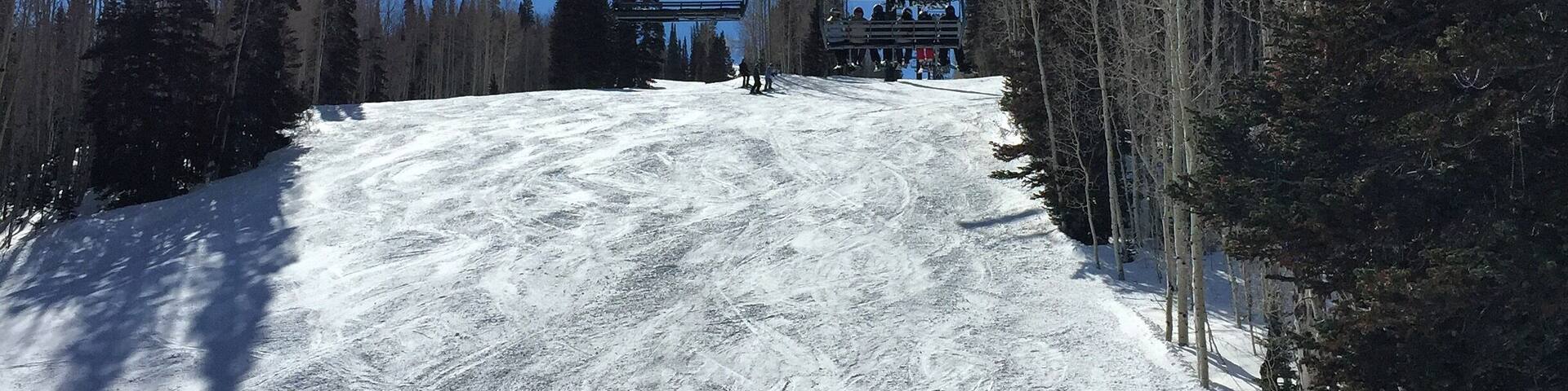 A ski lift with cables against a clear blue sky, surrounded by snow-covered slopes and tall evergreen trees.