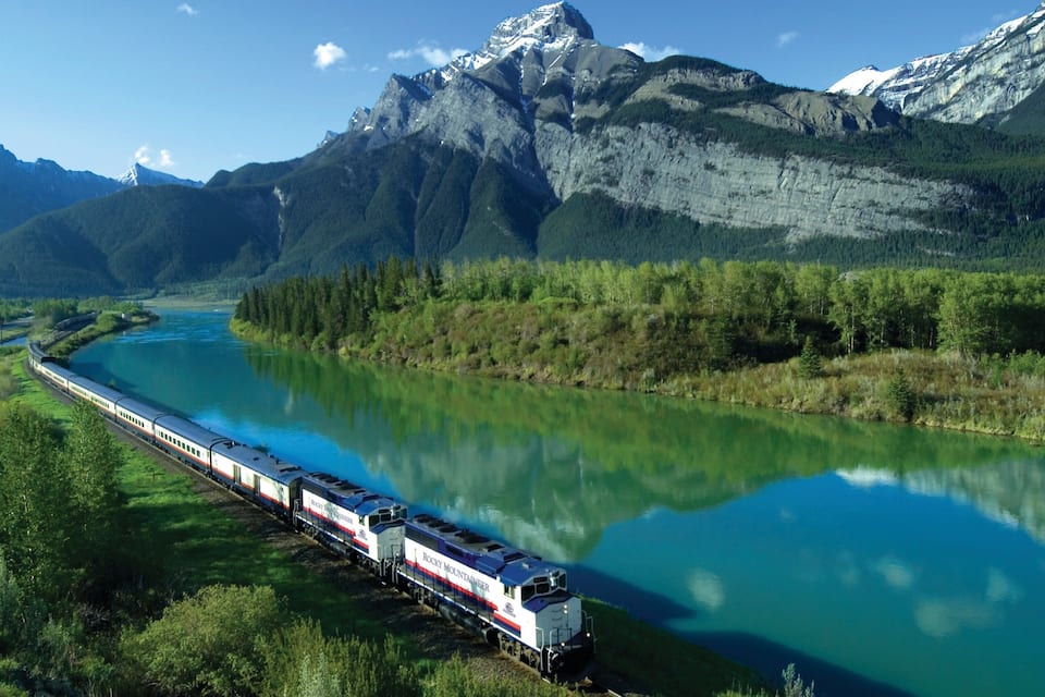 A Rocky Mountain Railway train traveling along a river surrounded by mountains.