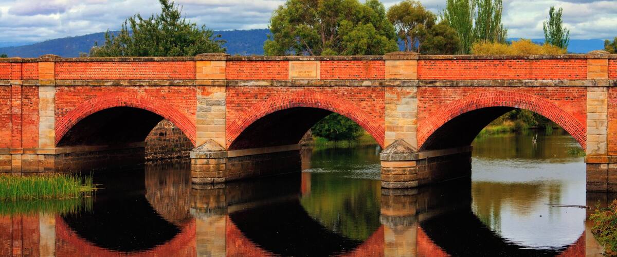 A red brick bridge with three arches over a calm river, surrounded by trees and hills.