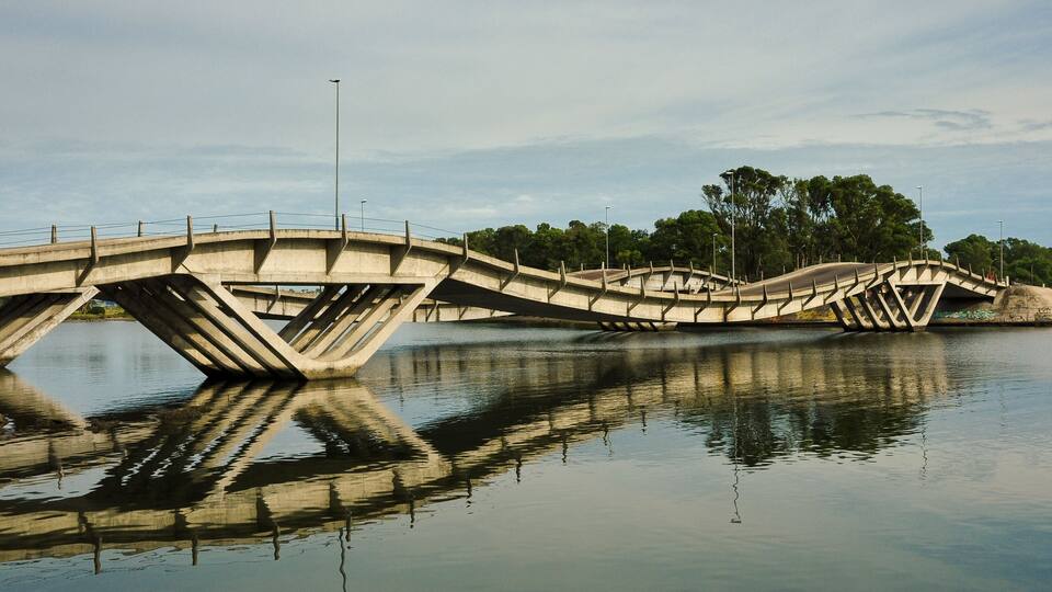 A concrete bridge with a unique design over a calm river, surrounded by trees.