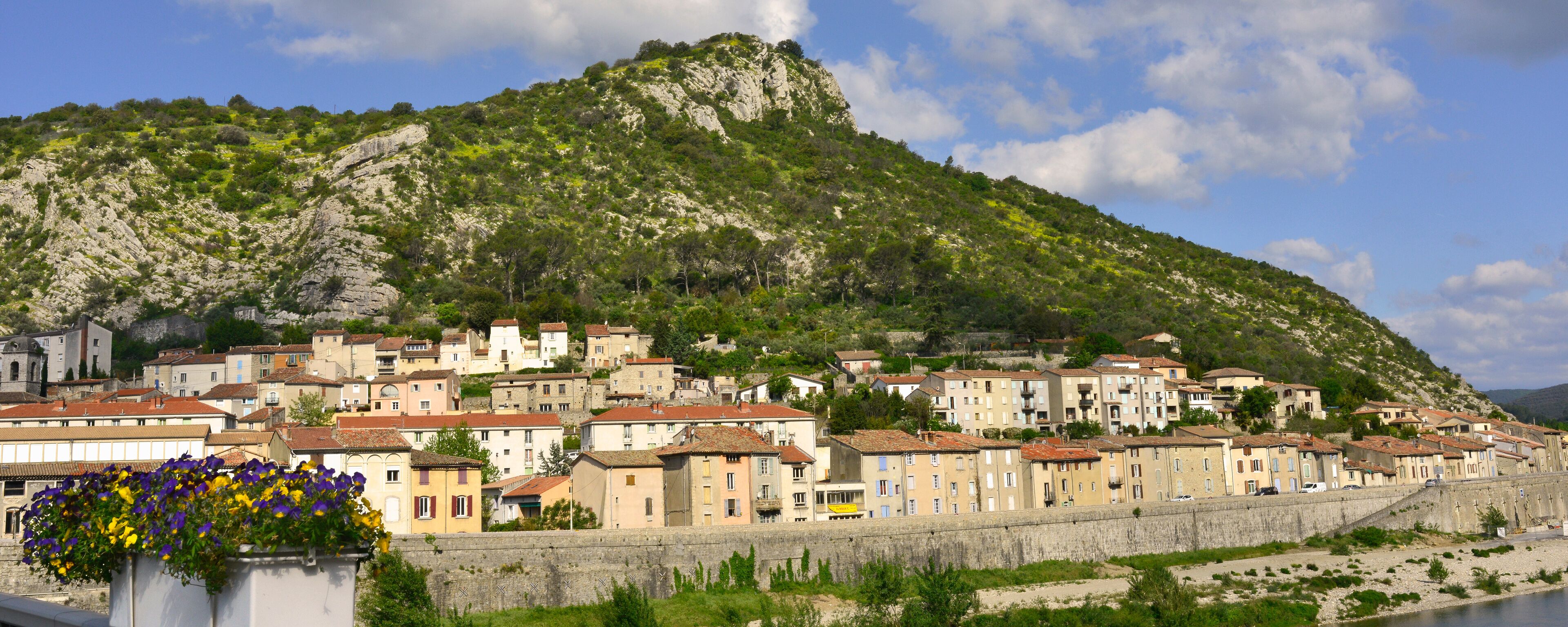 A coastal town with buildings along a stone wall, a hill with greenery, and a clear sky.