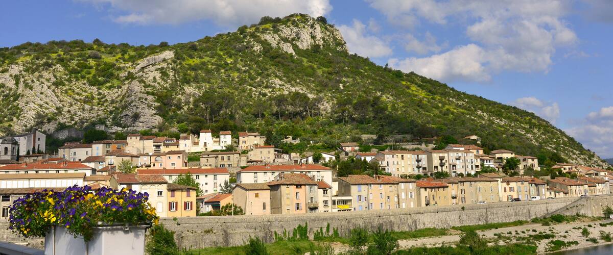 A coastal town with buildings along a stone wall, a hill with greenery, and a clear sky.