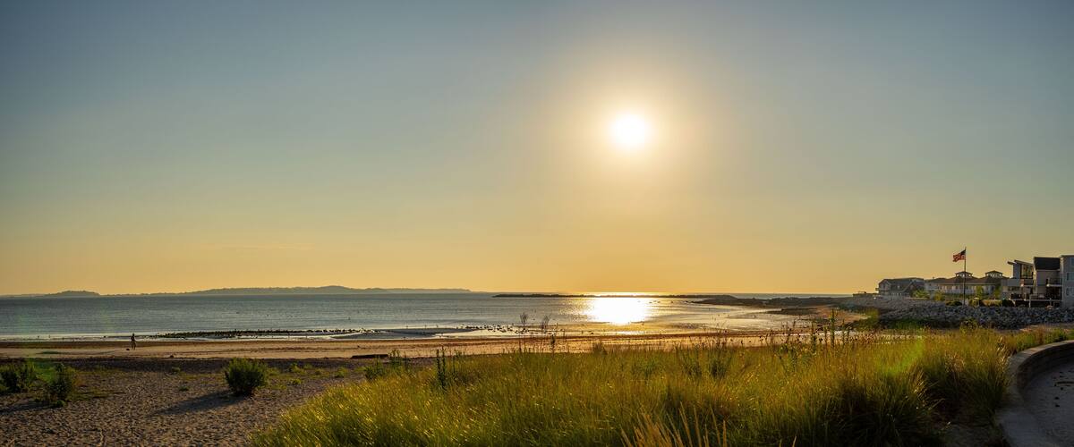A beach at sunset with the sun low on the horizon, a flag, and buildings in the background.