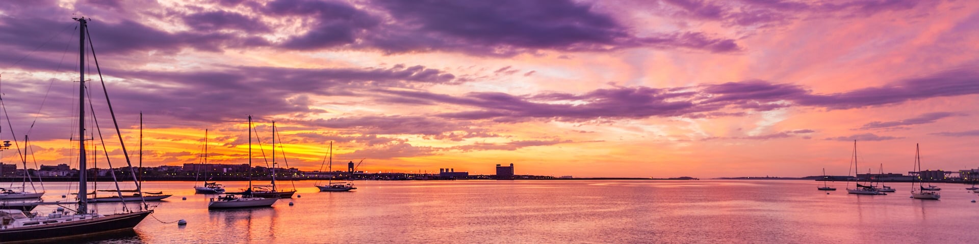 Um pôr do sol sobre um porto, com veleiros e a silhueta de uma cidade ao fundo.