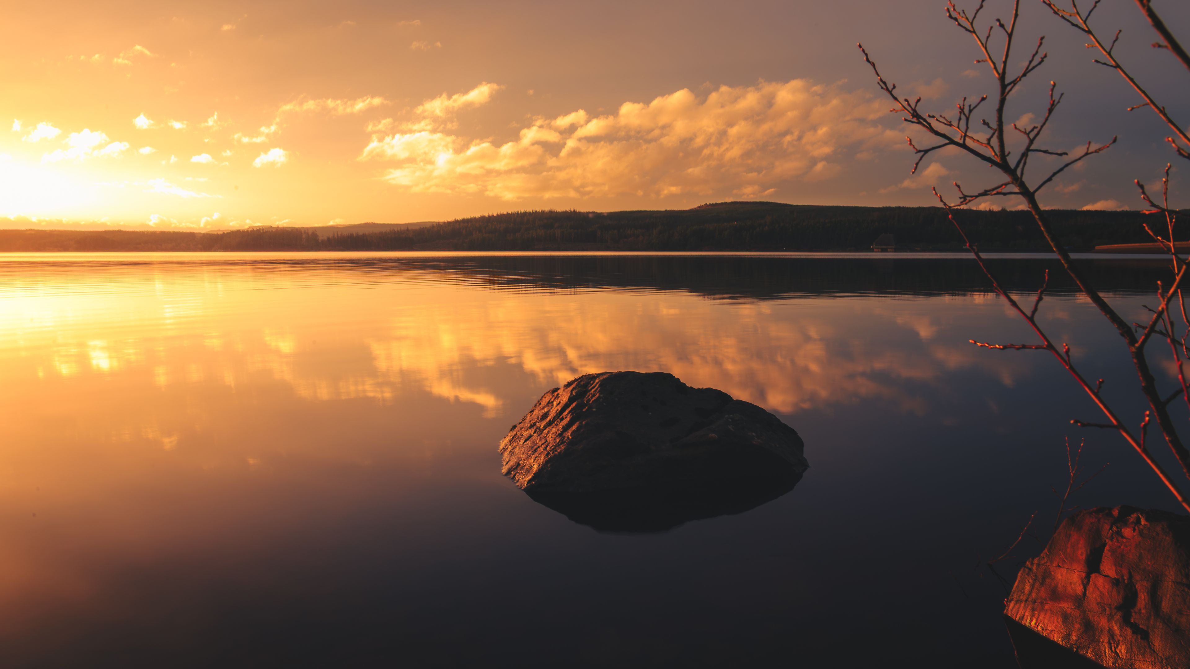 Un lago tranquilo al atardecer, con una gran roca en primer plano y una rama de árbol a la derecha.