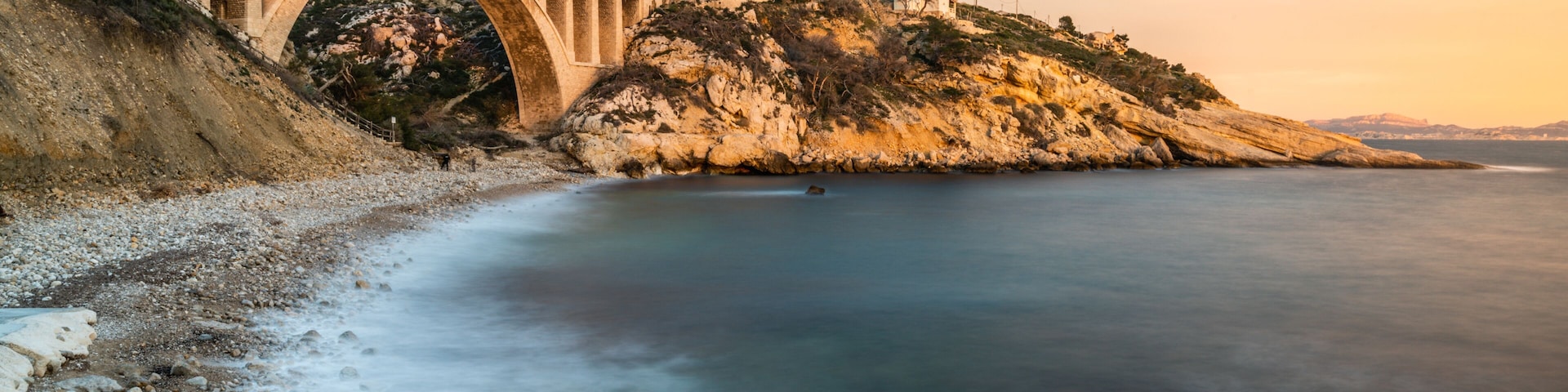 Un puente de piedra sobre un mar tranquilo, costa rocosa y una colina con vegetación escasa.