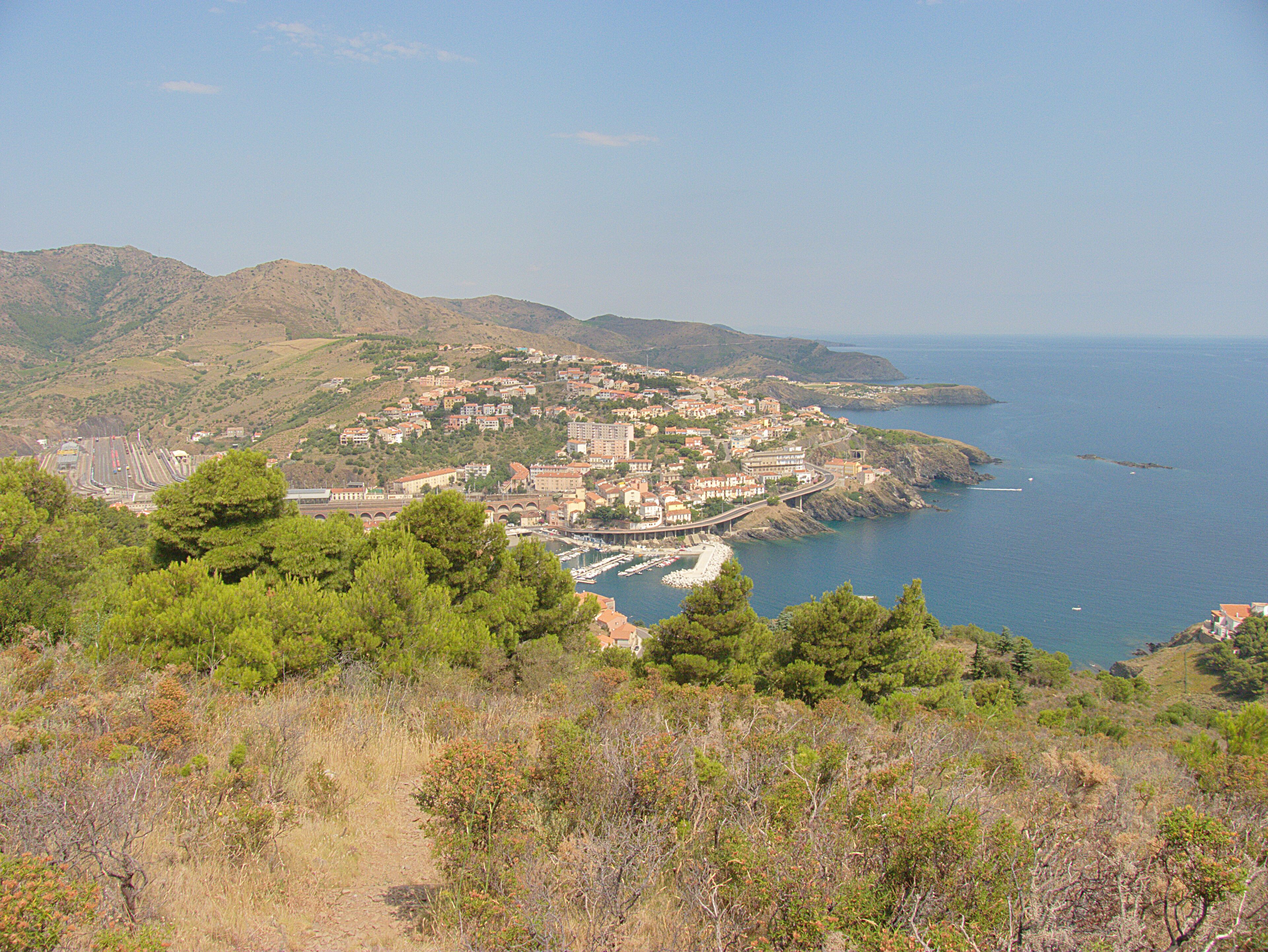 A coastal town with a marina, surrounded by hills and dense greenery.