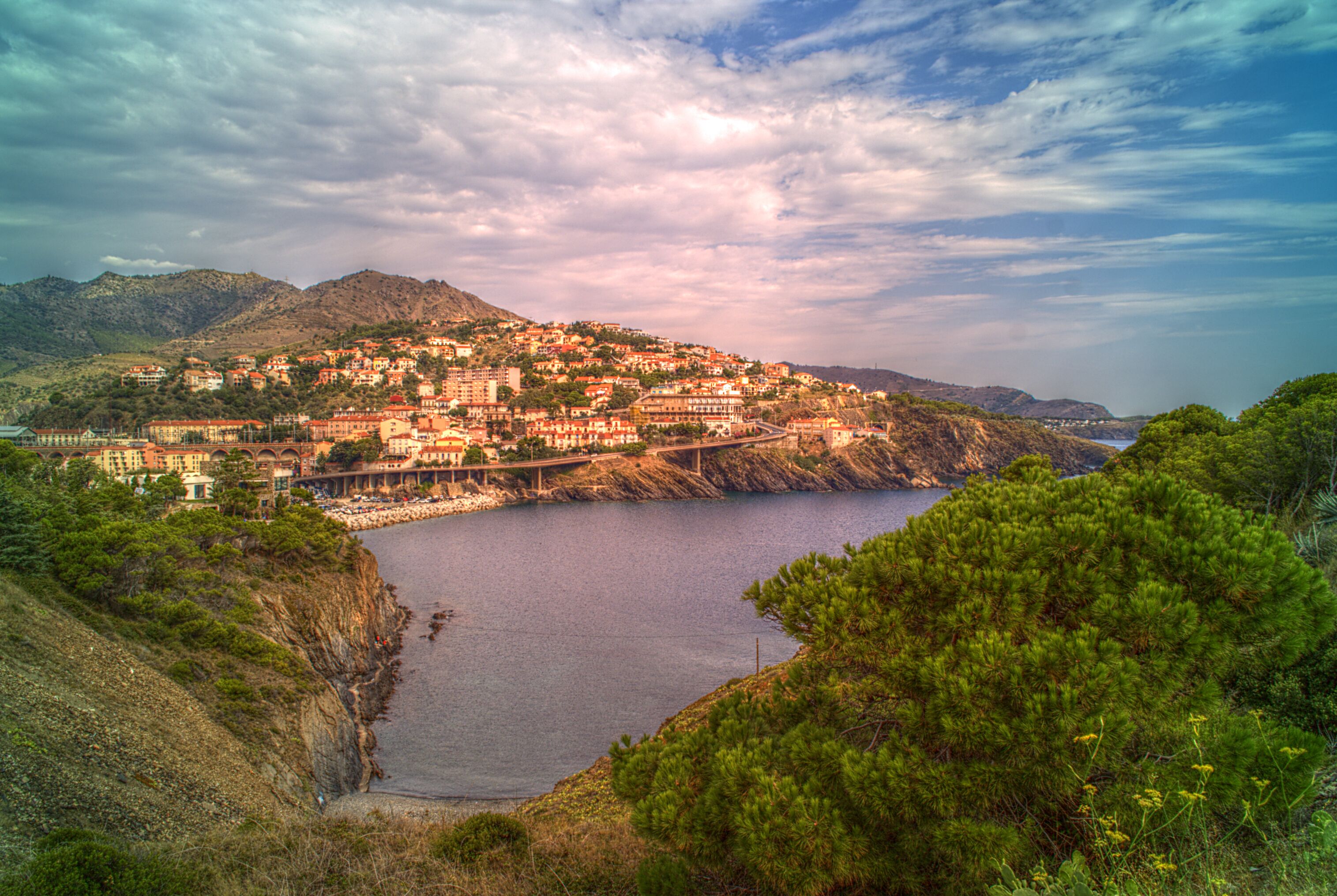 A coastal town with buildings on a hillside, a bridge, and a body of water.