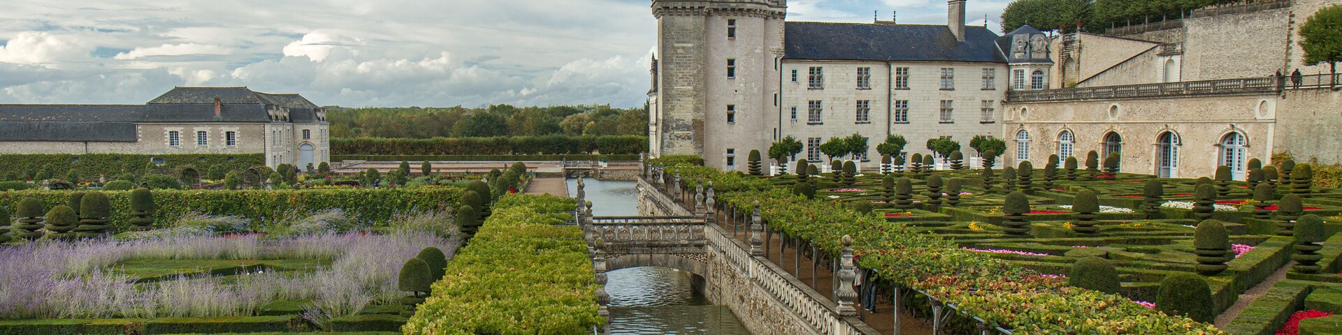 Un castillo histórico con un foso, jardines formales y un puente.