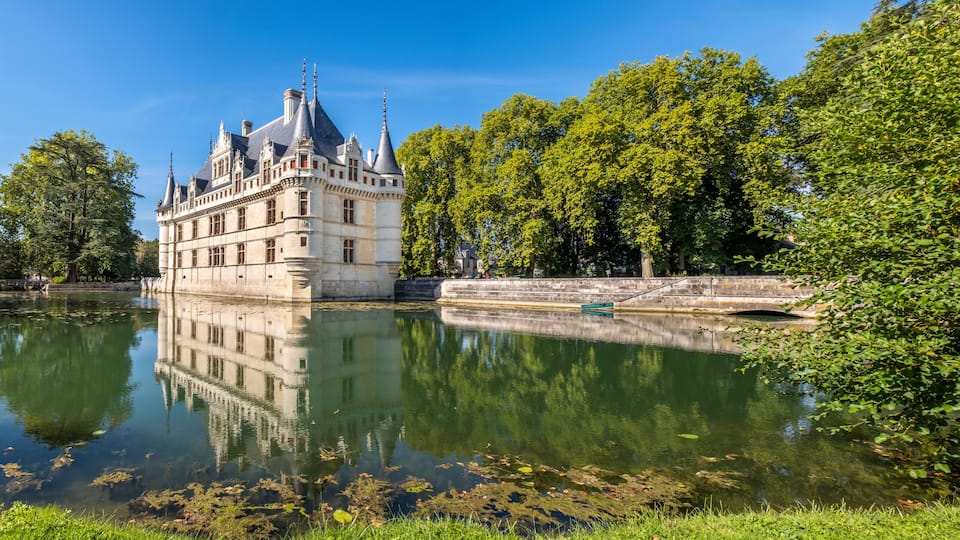 A historic château by a calm river, surrounded by lush greenery and a clear blue sky.