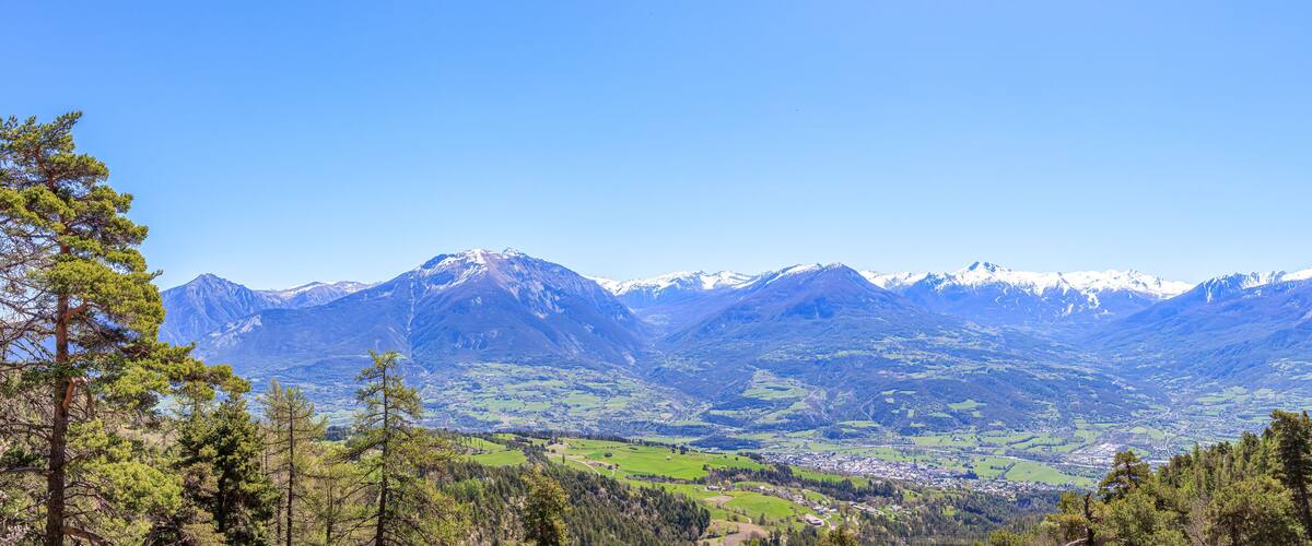 A scenic panoramic view of a mountain valley with a town surrounded by snowy mountain summit under a majestic blue sky