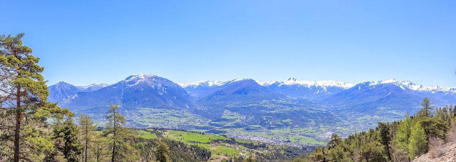 A scenic panoramic view of a mountain valley with a town surrounded by snowy mountain summit under a majestic blue sky
