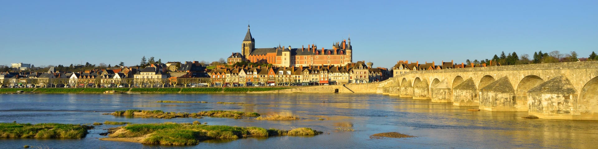 A historic building with a tower, a river with grassy patches, and a stone bridge.