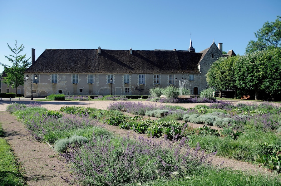 A historic stone building with a black roof, surrounded by a well-maintained garden with various plants and a paved walkway.