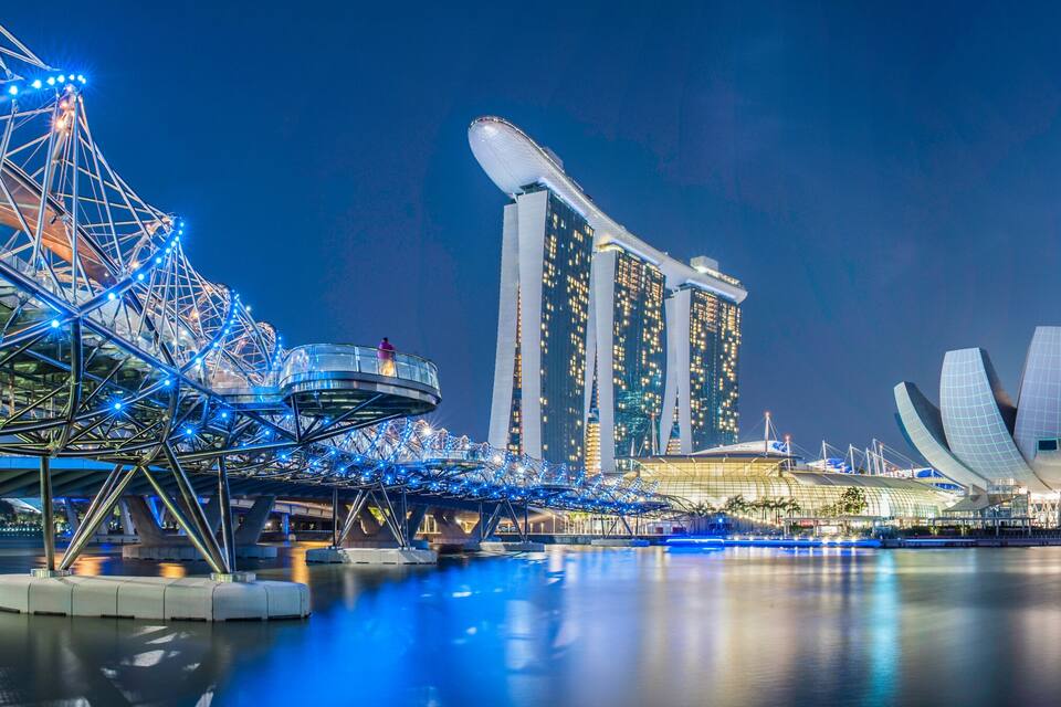 Uma paisagem urbana noturna com uma roda-gigante em destaque, um edifício moderno característico com telhado curvo e um conjunto de arranha-céus.
