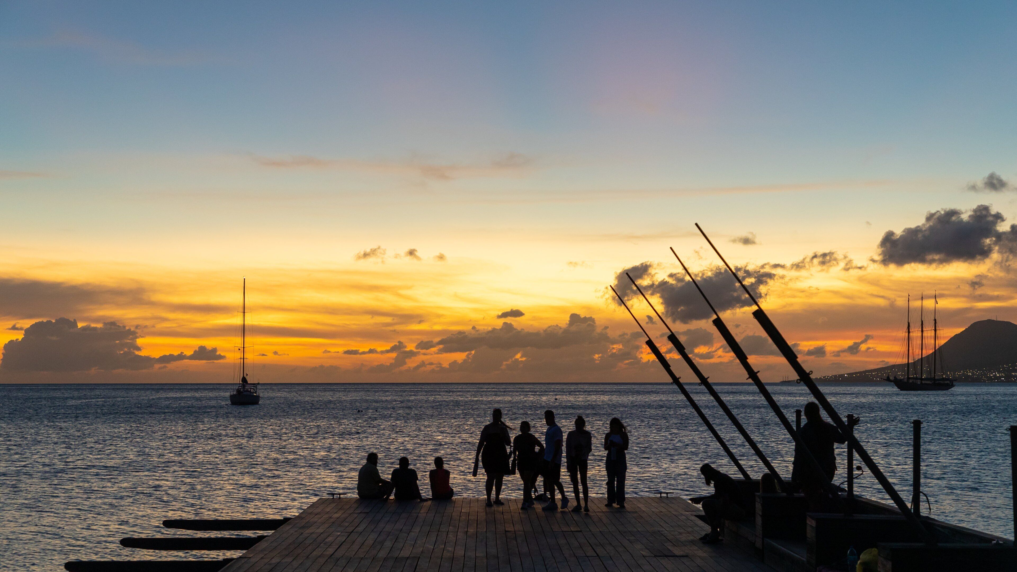 A sunset over a calm sea with people on a wooden pier, boats, and a distant mountain.