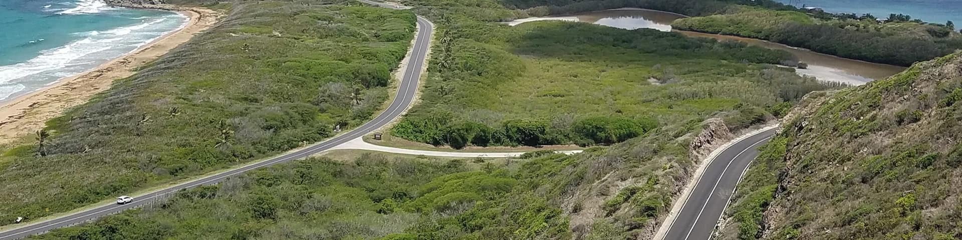 A coastal road winding through lush green hills with a beach and ocean in the background.
