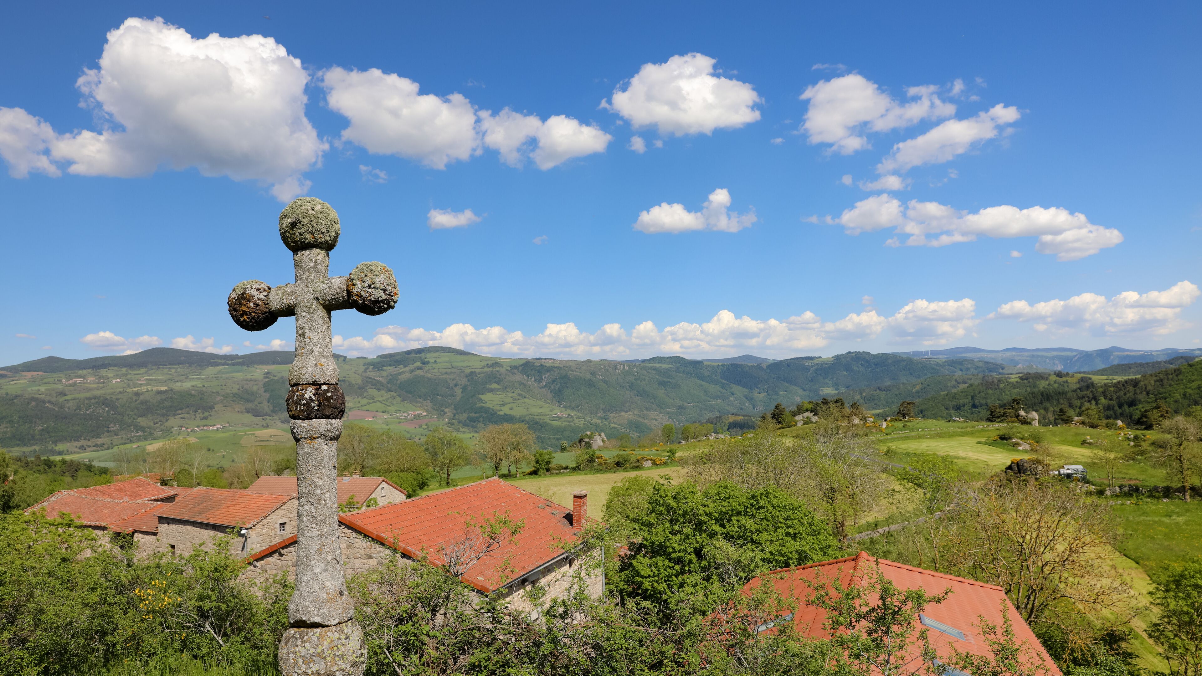 A stone cross in a rural landscape with rolling hills and red-roofed houses.
