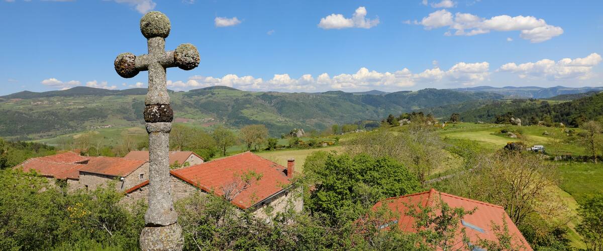 A stone cross in a rural landscape with rolling hills and red-roofed houses.