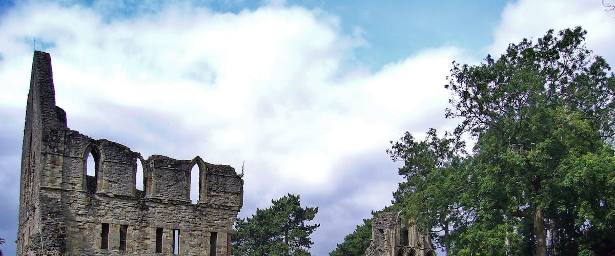 Les ruines d’une structure médiévale en pierre, dotée d’arches et de colonnes, sont entourées d’une végétation luxuriante et d’arbres.